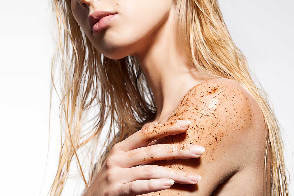 Woman applying textured exfoliating body scrub to her shoulder, illustrating the sensory experience of body care formulations.